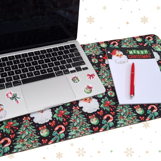A long rectangular desk mat with a festive Christmas print, styled on a white desk with a laptop, a notepad, and a red pen. The print features green pine trees, red berries, candy canes, ornaments, and Santa Claus motifs on a black background.
