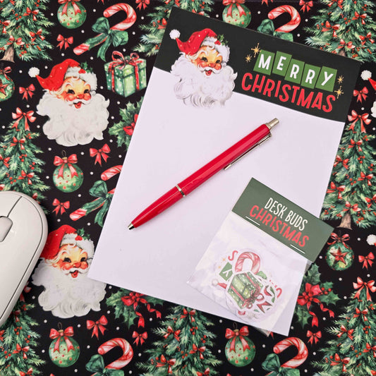 A styled flat-lay of Christmas-themed stationery on a festive patterned background. The set includes a white notepad with a black 'Merry Christmas' header and Santa illustration, a red pen, and a small pack of 'Desk Buds Christmas' stickers.
