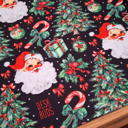 A close-up of a Christmas-themed desk mat featuring Santa Claus faces, candy canes, holly, ornaments, gift boxes, and the words 'DESK BUDS' on a black background.