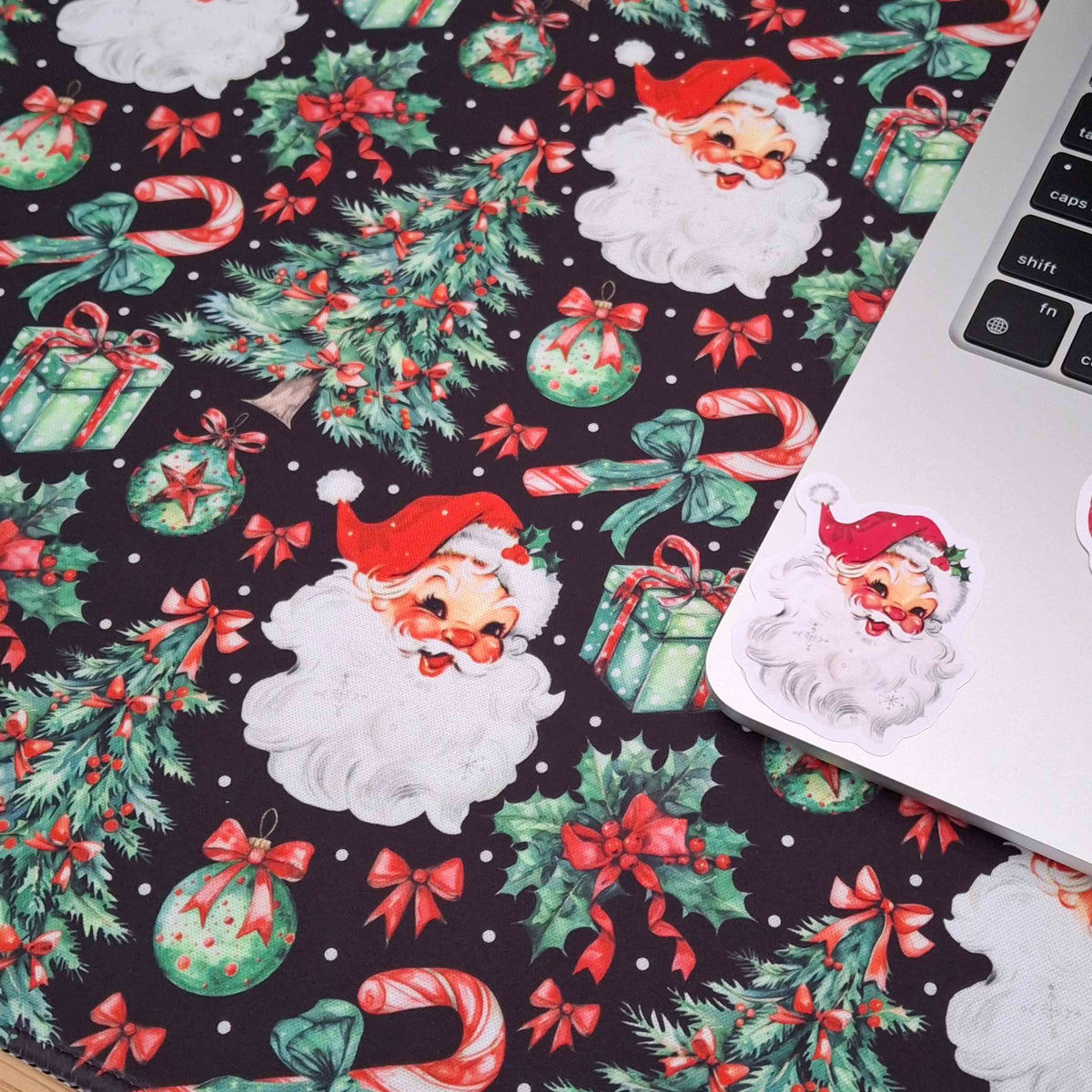 A close-up of a laptop resting on festive Christmas-themed extra large mouse pad. The paper features a repeating pattern of Santa Claus faces, green Christmas trees, candy canes, ornaments, and red bows on a dark background.