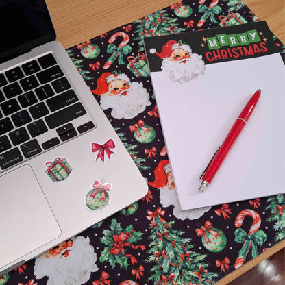 A festive Christmas-themed desk setup featuring a black mousepad with Santa Claus illustrations, a laptop, a notepad with a 'Merry Christmas' header, and a red pen.