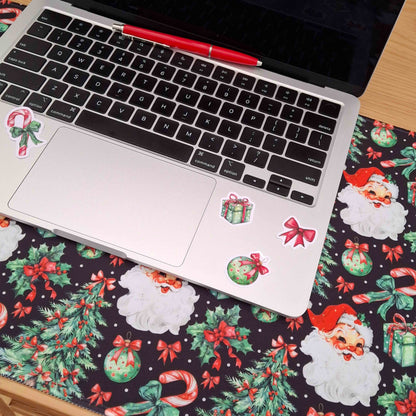A silver laptop sits on a dark Christmas-themed desk mat with Santa Claus, holly, candy canes, and red bows printed all over. A red pen rests on the keyboard.