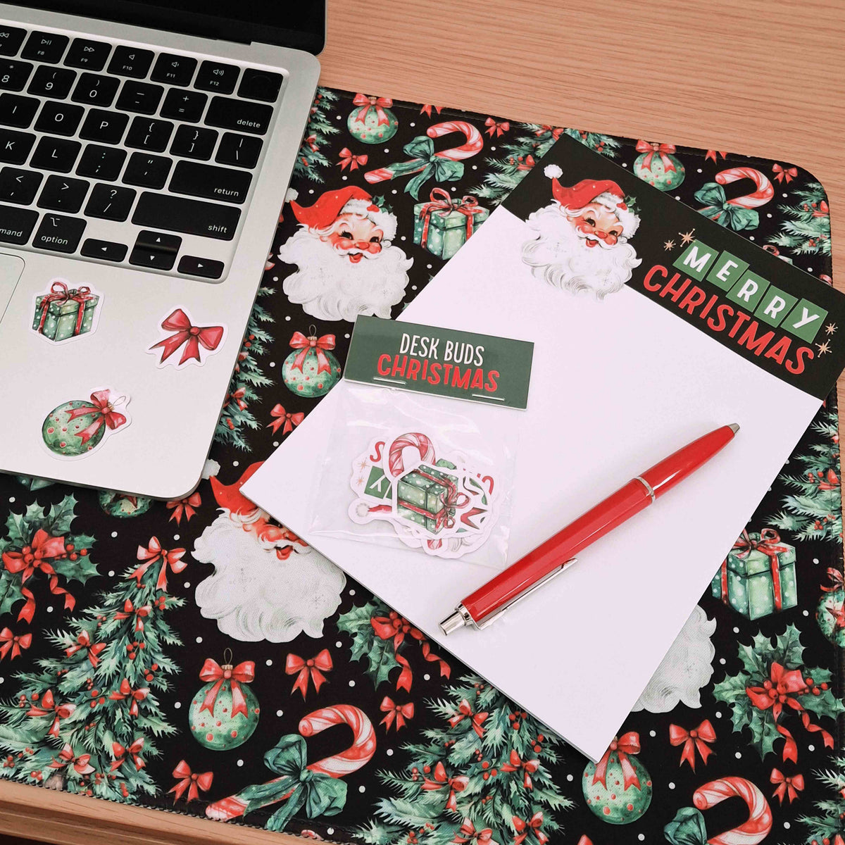 A Christmas-themed desk pad with a Santa and holiday pattern, styled with a laptop, a white notepad, a red pen, and a small sticker pack on a wooden desk.